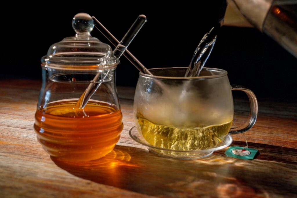 A steaming cup of tea being poured next to a jar of honey, set on a warm wooden table.